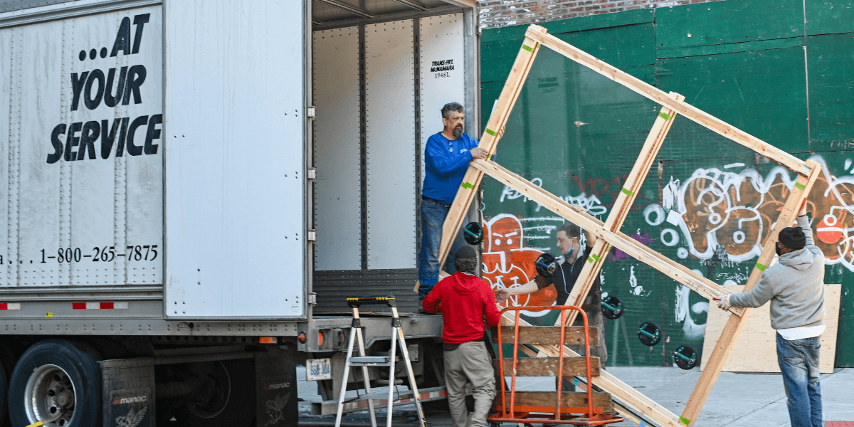 This image appears to show a group of people working together, unloading materials from a truck. They are handling a large wooden frame, possibly for a construction or installation project. The background includes a graffiti-covered wall and some tools, indicating an urban setting. The truck has a label on its side reading "...AT YOUR SERVICE" along with a contact number. The teamwork shown here suggests this might be part of a larger service or construction effort.