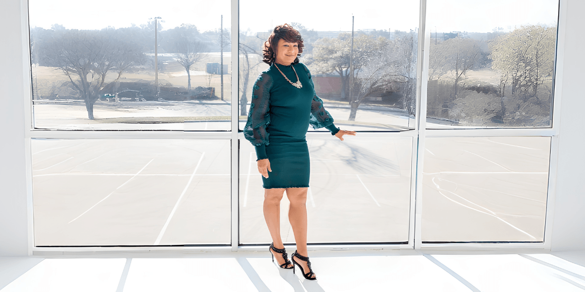 Woman in green dress posing by large window with sunlight, smiling confidently indoors.