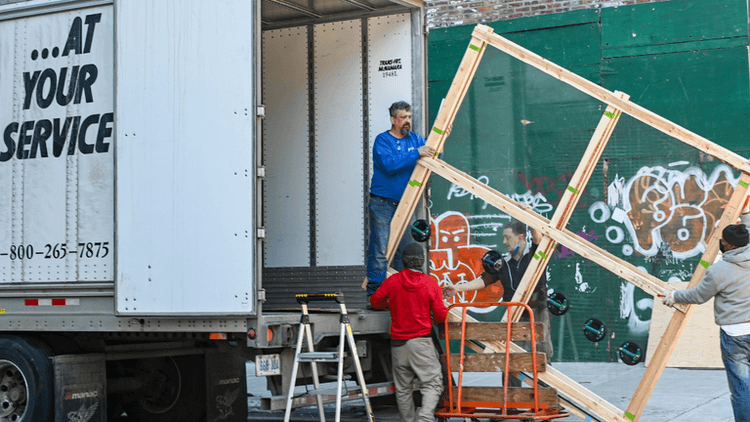 This image appears to show a group of people working together, unloading materials from a truck. They are handling a large wooden frame, possibly for a construction or installation project. The background includes a graffiti-covered wall and some tools, indicating an urban setting. The truck has a label on its side reading "...AT YOUR SERVICE" along with a contact number. The teamwork shown here suggests this might be part of a larger service or construction effort.