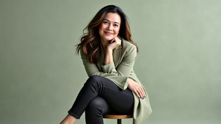 A woman with shoulder-length hair, wearing a green blazer, sitting on a stool, smiling confidently in a calm setting.