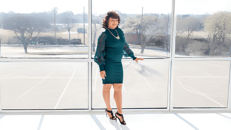 Woman in green dress posing by large window with sunlight, smiling confidently indoors.