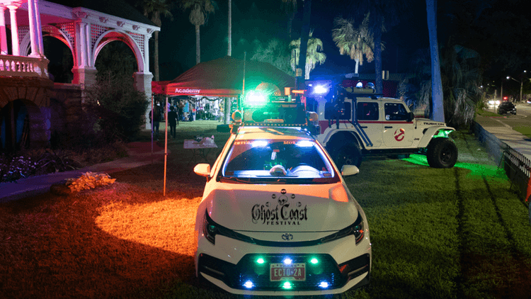 A Toyota car and Ghostbusters-themed Jeep at the Ghost Coast Festival, showcasing a lively, family-friendly atmosphere with vibrant lighting.