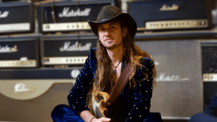 Rock guitarist with long hair and cowboy hat sitting with electric guitar in front of stacked Marshall amplifiers in a music studio