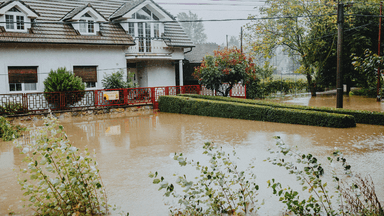 Flooded residential area with water surrounding a house and garden under overcast skies