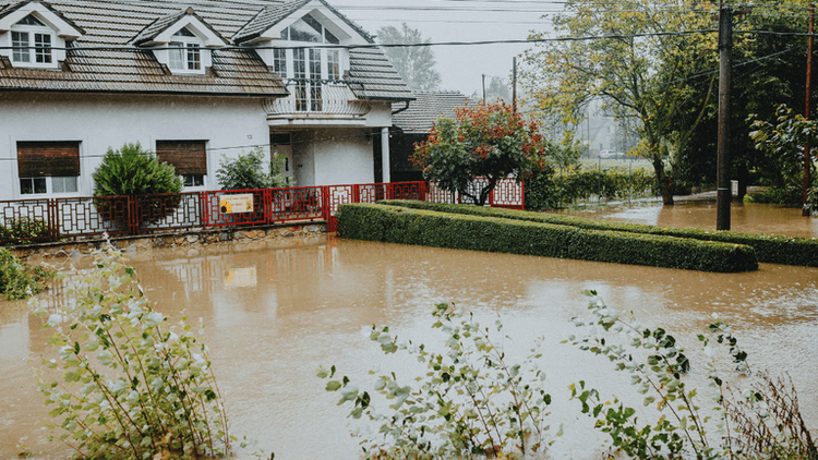 Flooded residential area with water surrounding a house and garden under overcast skies