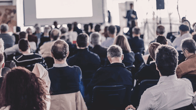 Audience seated in a conference room, listening to a presenter on stage