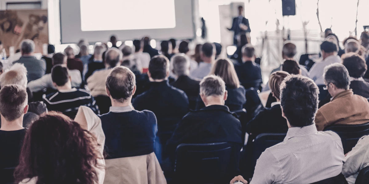 Audience seated in a conference room, listening to a presenter on stage