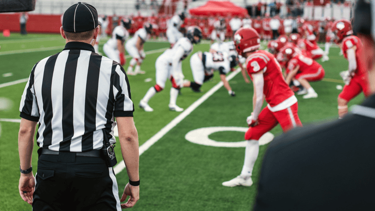 A referee observes two football teams lined up on the field ready for play
