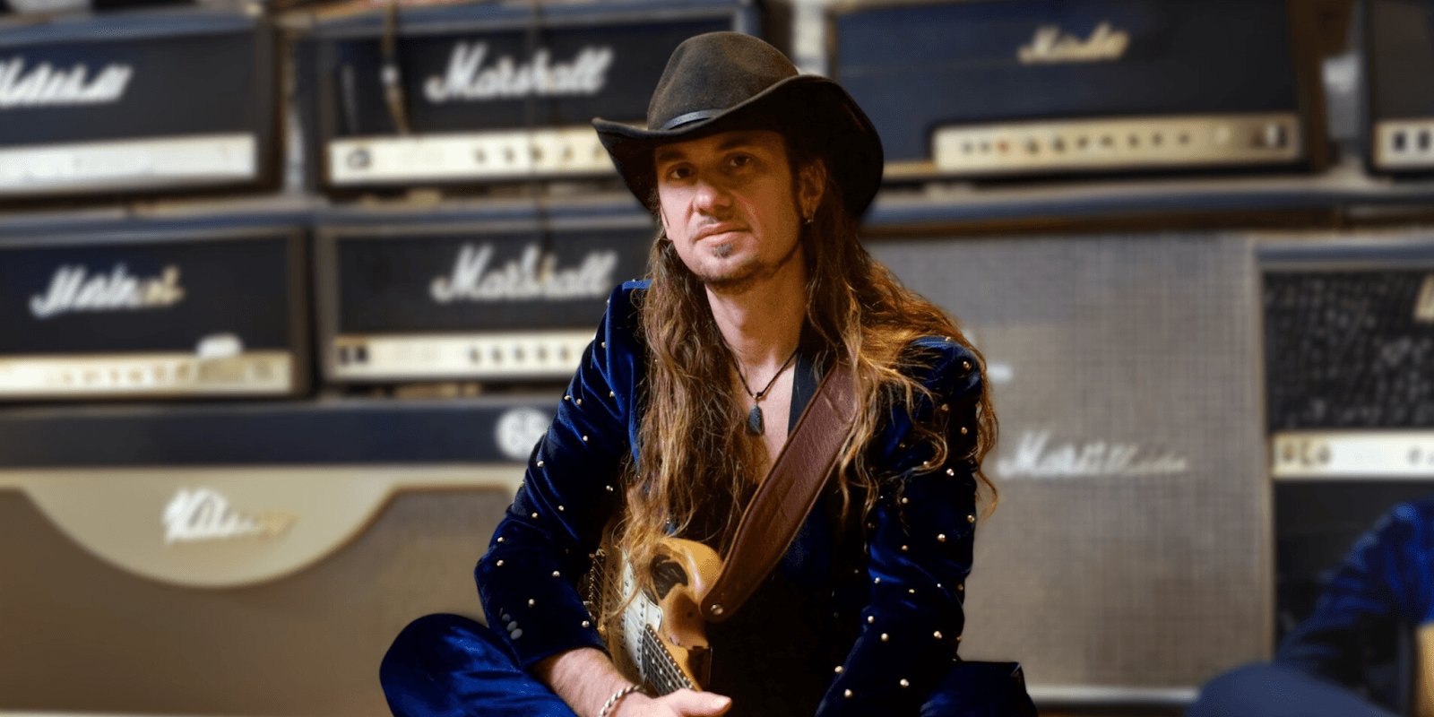 Rock guitarist with long hair and cowboy hat sitting with electric guitar in front of stacked Marshall amplifiers in a music studio