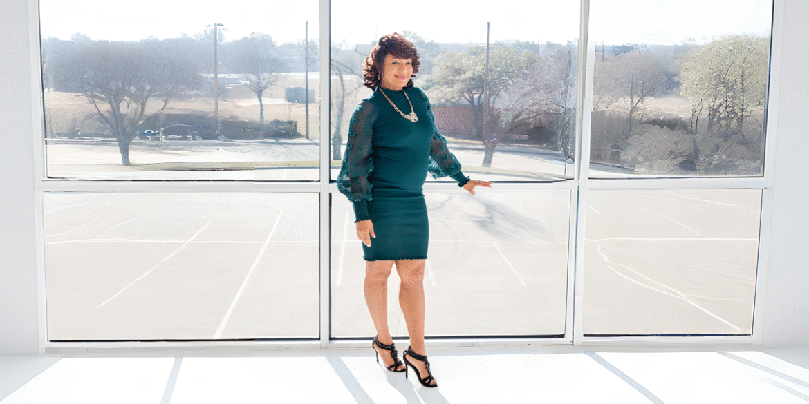 Woman in green dress posing by large window with sunlight, smiling confidently indoors.